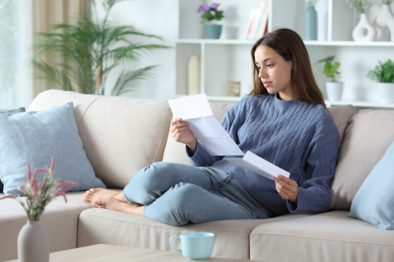 Woman sitting on a sofa in a bright living room, reading a letter and holding an envelope, with a coffee cup on the table nearby.