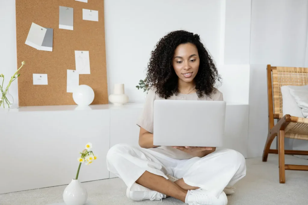 Woman sitting on the floor in a bright workspace, using a laptop with mood-board notes and minimal décor behind her.
