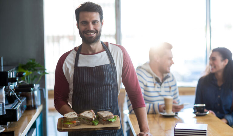 Smiling café worker in a striped apron holds a wooden board of sandwiches beside a counter, with customers chatting and coffee and pastries in the background.