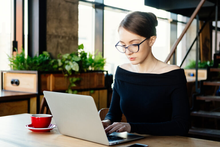 Woman wearing glasses works on a laptop at a café table, with a red coffee cup beside her and greenery and large windows in the background.