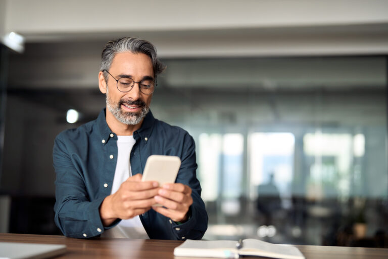Man with glasses checks his phone at a desk, with an open notebook in front of him and a blurred office background.
