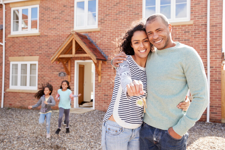 Couple smiles outside a brick house, holding up new keys, while two children run toward them from the open front door.