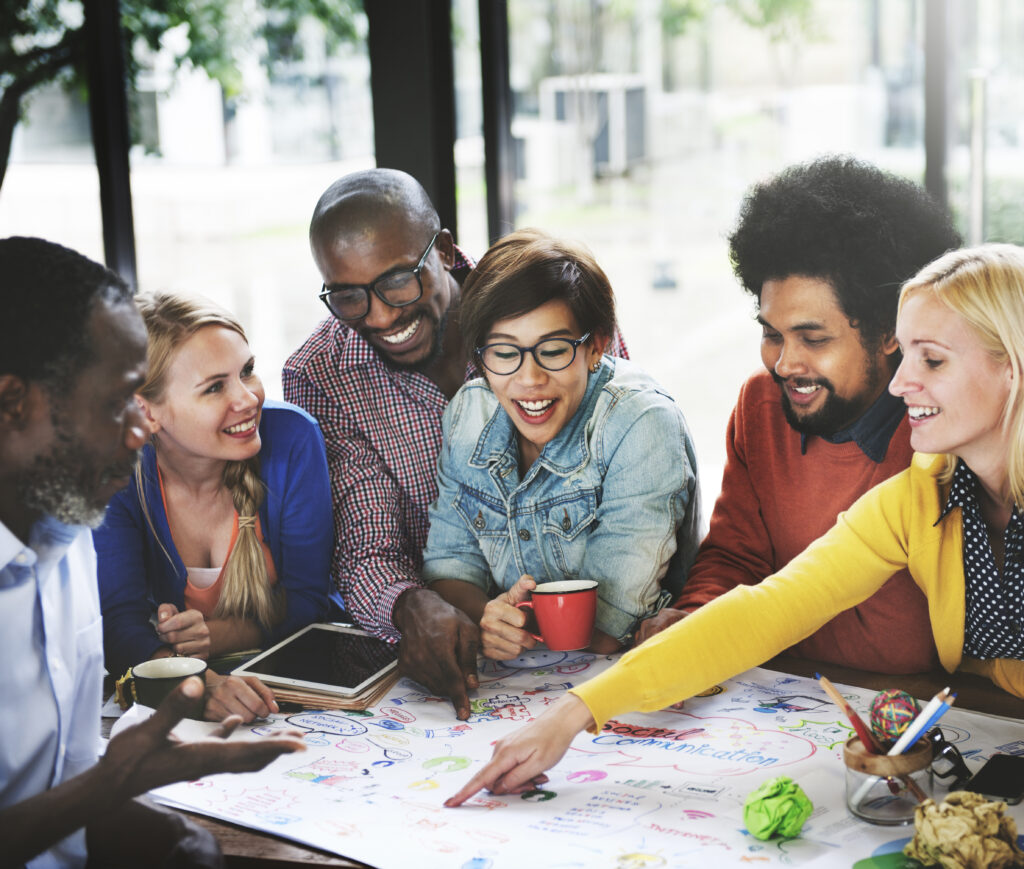Group of adult learners gather around a table, smiling and pointing at a colorful brainstorming sheet with notes, pens, and coffee mugs.
